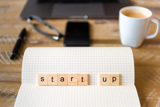 Closeup on notebook over wood table background, focus on wooden blocks with letters making Start Up word. Business concept image. Laptop, glasses, pen and mobile phone in a defocused background.
