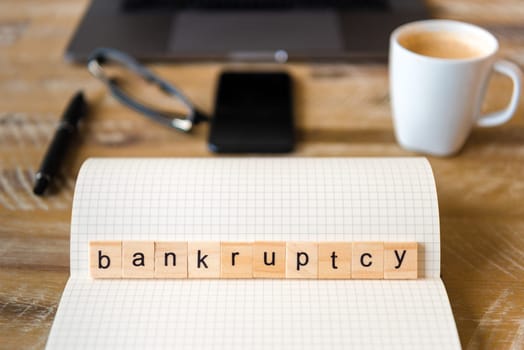 Closeup on notebook over wood table background, focus on wooden blocks with letters making Bankruptcy word. Business concept image. Laptop, glasses, pen and mobile phone in a defocused background.