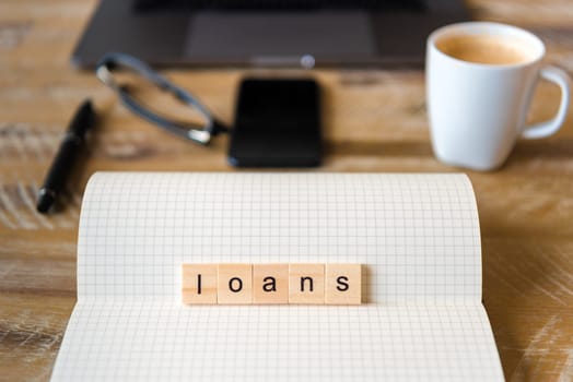 Closeup on notebook over wood table background, focus on wooden blocks with letters making Loans word. Business concept image. Laptop, glasses, pen and mobile phone in a defocused background.