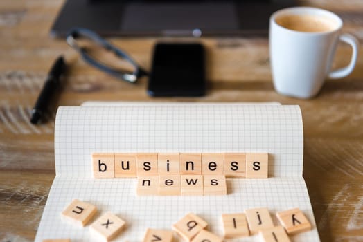 Closeup on notebook over wood table background, focus on wooden blocks with letters making Business News words. Business concept image. Laptop, glasses, pen and mobile phone in a defocused background.