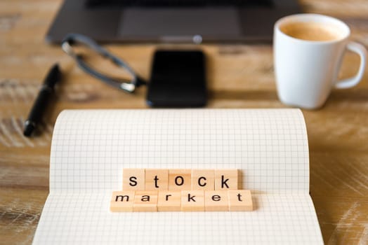 Closeup on notebook over wood table background, focus on wooden blocks with letters making Stock Market words. Business concept image. Laptop, glasses, pen and mobile phone in a defocused background.