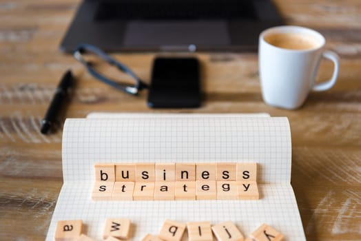Closeup on notebook over wood table background, focus on wooden blocks with letters making Business Strategy words. Concept image with laptop, glasses, pen and mobile phone in a defocused background.