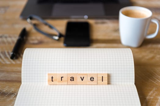 Closeup on notebook over wood table background, focus on wooden blocks with letters making Travel word. Business concept image. Laptop, glasses, pen and mobile phone in a defocused background.