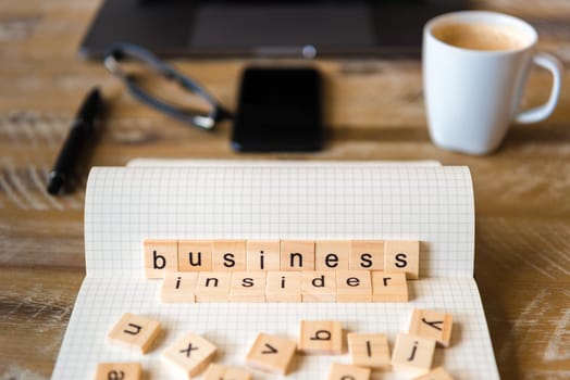 Closeup on notebook over wood table background, focus on wooden blocks with letters making Business Insider words. Business concept image. Laptop, glasses, pen and mobile phone in defocused background