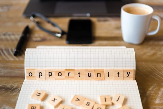 Closeup on notebook over wood table background, focus on wooden blocks with letters making Opportunity word. Business concept image. Laptop, glasses, pen and mobile phone in a defocused background.