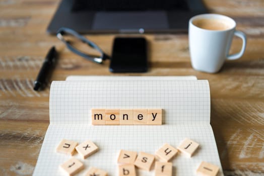 Closeup on notebook over wood table background, focus on wooden blocks with letters making Money word. Business concept image. Laptop, glasses, pen and mobile phone in a defocused background.