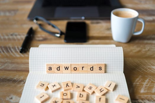 Closeup on notebook over wood table background, focus on wooden blocks with letters making Adwords word. Business concept image. Laptop, glasses, pen and mobile phone in a defocused background.