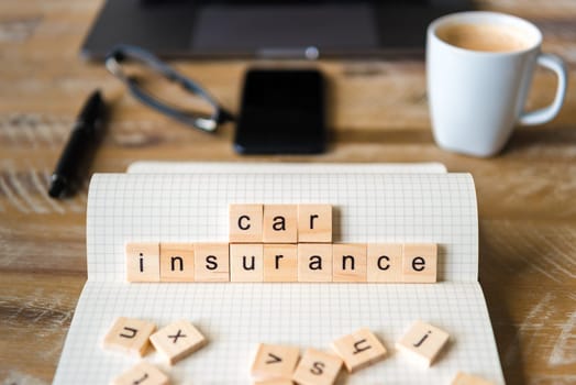 Closeup on notebook over wood table background, focus on wooden blocks with letters making Car Insurance words. Business concept image. Laptop, glasses, pen and mobile phone in a defocused background.