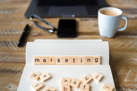 Closeup on notebook over wood table background, focus on wooden blocks with letters making Marketing word. Business concept image. Laptop, glasses, pen and mobile phone in a defocused background.