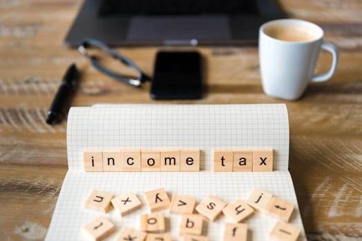 Closeup on notebook over wood table background, focus on wooden blocks with letters making Income Tax words. Business concept image. Laptop, glasses, pen and mobile phone in a defocused background.