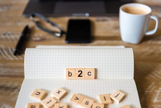 Closeup on notebook over wood table background, focus on wooden blocks with letters making B2C word. Business concept image. Laptop, glasses, pen and mobile phone in a defocused background.