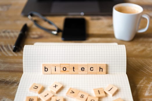 Closeup on notebook over wood table background, focus on wooden blocks with letters making Biotech word. Business concept image. Laptop, glasses, pen and mobile phone in a defocused background.