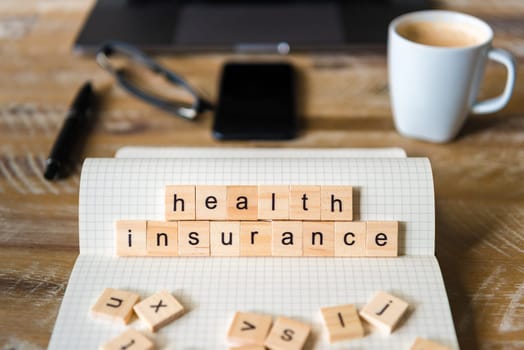 Closeup on notebook over wood table background, focus on wooden blocks with letters making Health Insurance words. Concept image with laptop, glasses, pen and mobile phone in a defocused background.