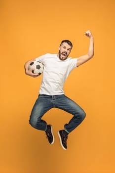 The young man as soccer football player jumping and kicking the ball at studio on a yellow background. Jumping footballer in a combat kick. Football fan and world championship concept. Human emotions