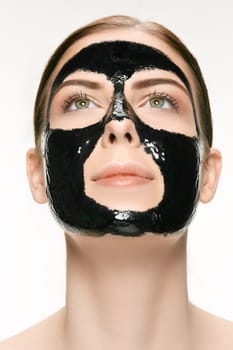 Young beautiful caucasian woman applying a black cosmetic mask for the face of the therapeutic black mud. Spa treatment and face beauty concept. Female face care and perfect skin concepts