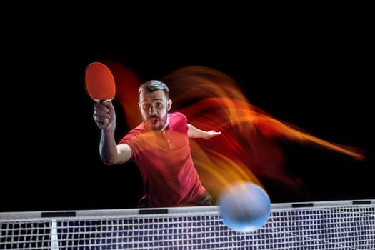 The table tennis player in motion. Fit young sports man tennis-player in play on black background with lights. Movement, sport game, stobe concepts. Professional. Human emotions, facial expression