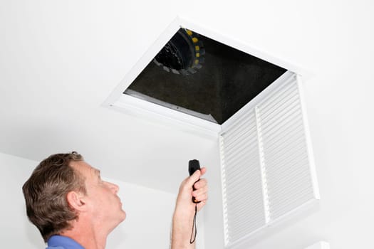 Male looking up into a ceiling air intake duct with a flashlight checking for maintenance. Person with a flashlight examining with a square opening of a home HVAC system.