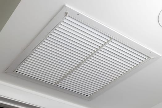 One large white painted metal furnace air vent grill with many openings on a ceiling close-up. Square outflow air filter door vent in a modern home ceiling up close.