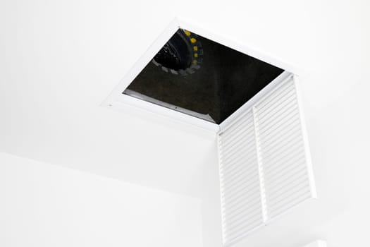 White square ceiling furnace air vent grate open, showing inside the duct. Metal air intake grate open and hanging from the ceiling showing view of inside the HVAC system.