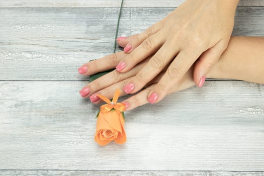 Hands of woman pink manicured on nails and an beautiful orange rose . Background is a rustic bright table. Concept beauty.