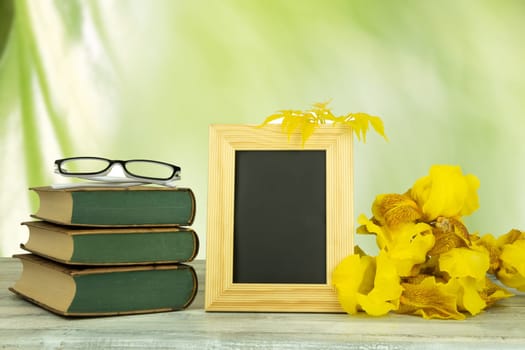 Empty frame with a bouquet of yellow flowers and a pair of glasses on old books over bright green natural background. Back to school concept. The teachers day.