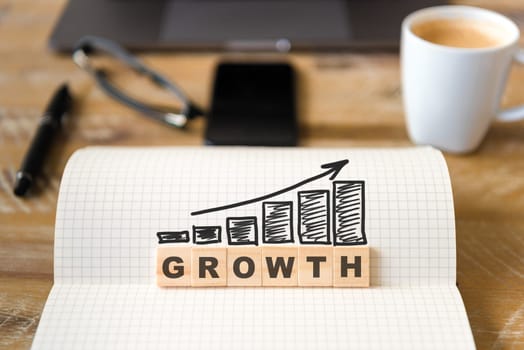Closeup on notebook over wood table background, focus on wooden blocks with letters making GROWTH word and rising arrow over graph bars. Laptop, glasses, pen and mobile phone in defocused background