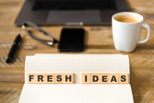 Closeup on notebook over wood table background, focus on wooden blocks with letters making Fresh Ideas text. Concept image with laptop, glasses, pen and mobile phone in defocused background