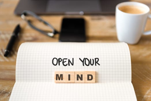Closeup on notebook over wood table background, focus on wooden blocks with letters making Open Your Mind text. Concept image with laptop, glasses, pen and mobile phone in defocused background