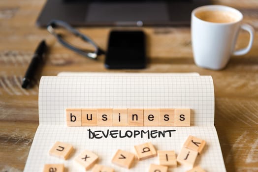 Closeup on notebook over wood table background, focus on wooden blocks with letters making Business Development text. Concept image with laptop, glasses, pen and mobile phone in defocused background
