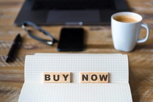 Closeup on notebook over wood table background, focus on wooden blocks with letters making Buy Now text. Concept image with laptop, glasses, pen and mobile phone in defocused background