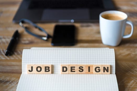 Closeup on notebook over wood table background, focus on wooden blocks with letters making JOB DESIGN words. Business concept image. Laptop, glasses, pen and mobile phone in defocused background