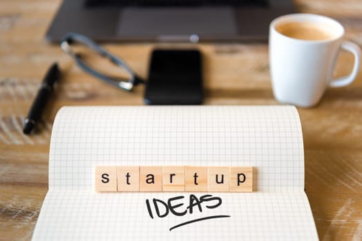 Closeup on notebook over wood table background, focus on wooden blocks with letters making Startup Ideas text. Concept image with laptop, glasses, pen and mobile phone in defocused background