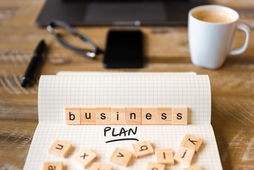 Closeup on notebook over wood table background, focus on wooden blocks with letters making Business Plan text. Concept image with laptop, glasses, pen and mobile phone in defocused background