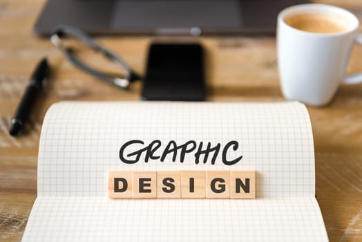 Closeup on notebook over wood table background, focus on wooden blocks with letters making Graphic Design text. Concept image with laptop, glasses, pen and mobile phone in defocused background