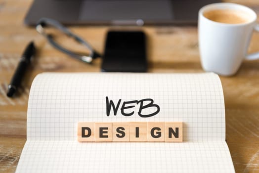 Closeup on notebook over wood table background, focus on wooden blocks with letters making Web Design text. Concept image with laptop, glasses, pen and mobile phone in defocused background