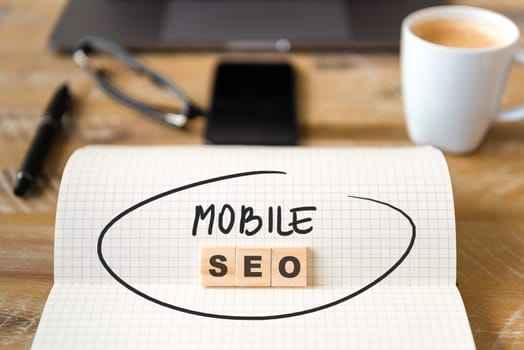 Closeup on notebook over wood table background, focus on wooden blocks with letters making Mobile SEO text. Business concept image. Laptop, glasses, pen and mobile phone in defocused background