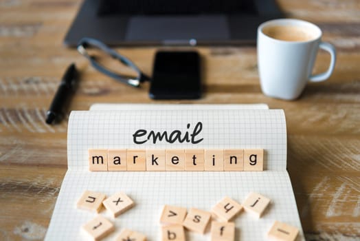 Closeup on notebook over wood table background, focus on wooden blocks with letters making Email Marketing text. Concept image with laptop, glasses, pen and mobile phone in defocused background