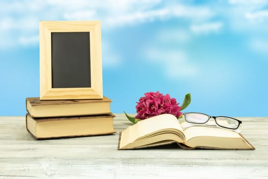 Empty frame on old books with a blossom from pink azalee and a pair of glasses on old book over bright blue sky background. Back to school concept. The teachers day.