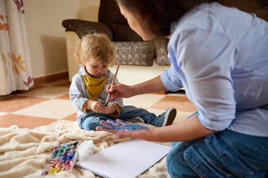 A mother and her young child bond through an artistic activity, utilizing paints as they share a creative session on a comfortable blanket in their living room.
