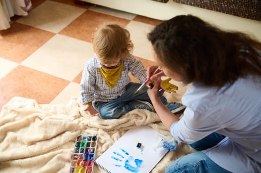 A mother engages with her child in a creative painting activity, using watercolors to paint on paper and their hands, depicting joyful moments and parental bonding.
