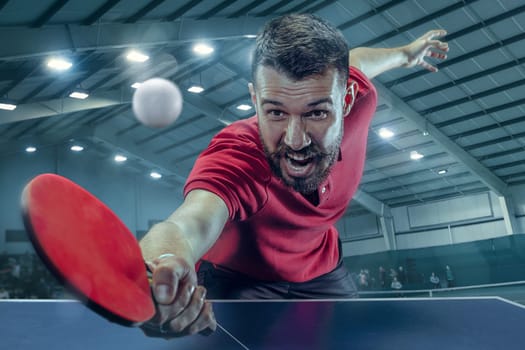 The table tennis player in motion. Fit young sports man tennis-player in play on sport arena background with lights. Movement, sport game, stobe concepts. Professional. Human emotions, facial expression