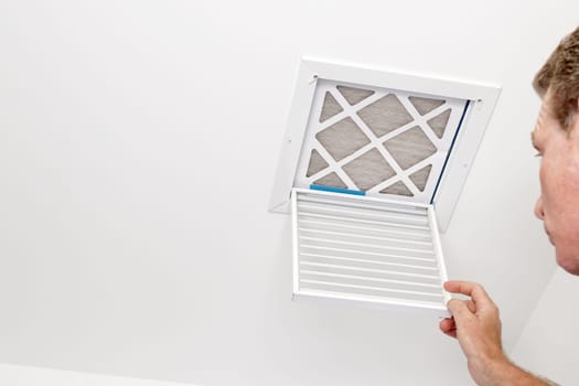 Mature adult opening a small square HVAC air intake vent revealing a dirty air filter in the ceiling. Man opened a a vent grill cover to a dirty ceiling air filter..