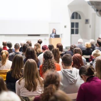 Female speaker giving presentation in lecture hall at university workshop. Audience in conference hall. Rear view of unrecognized participant in audience. Scientific conference event.