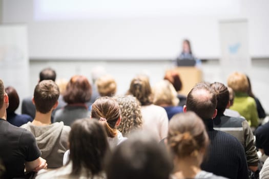 Female speaker giving presentation in lecture hall at university workshop. Audience in conference hall. Rear view of unrecognized participant in audience. Scientific conference event.