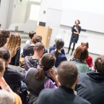 Female speaker giving presentation in lecture hall at university workshop. Audience in conference hall. Rear view of unrecognized participant in audience. Scientific conference event.