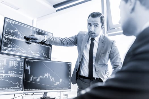 Businessmen trading stocks online. Stock brokers looking at graphs, indexes and numbers on multiple computer screens. Colleagues in discussion in traders office. Blue toned black and white.