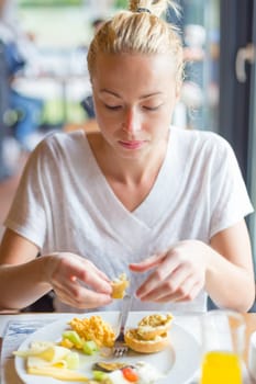 Female tourist eating delicious healthy breakfast at one of street cafes: scrambled eggs, cheese, vegetables, bread and orange juice.