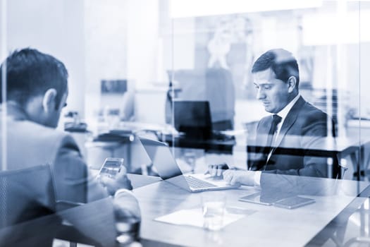 Through the window view of two young businessmen using laptop computer and smart phone devices at working business meeting in corporative office. Black and white blue toned image.
