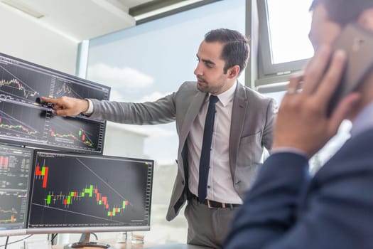Businessmen trading stocks online. Stock brokers looking at graphs, indexes and numbers on multiple computer screens. Colleagues in discussion in traders office. Business success concept.