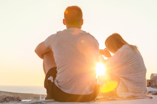 Rear view of unrecognizable young couple watching sunrise and taking vacation photos at Santorini island, Greece.